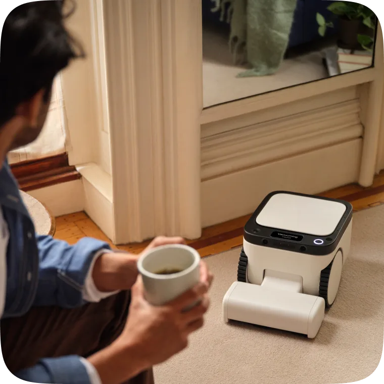 Matic robot vacuuming a living-room carpet beside a person holding coffee